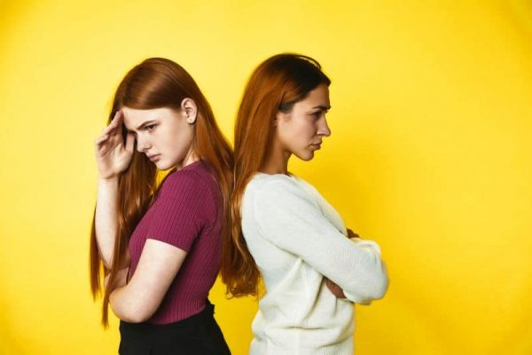 Two upset redhead caucasian girls are standing disappointed back to back, on the yellow background dressed in casual clothes