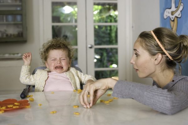 Toddler struggling and crying in high chair while woman picks up goldfish snacksat the dining table.
